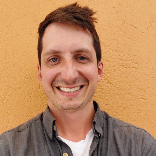 Man with orange backdrop, smiling looking straight ahead. Wearing grey collared shirt. 