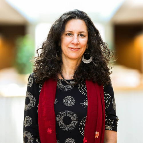 Woman with dark curly hair smiling. Wearing patterned shirt with red scarf.