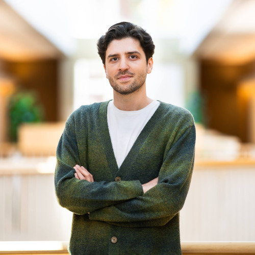 Man with dark hair standing with his arms crossed. He is wearing a green cardigan and white shirt.
