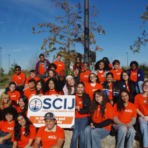 Group of students in orange shirts smiling with a blue sky behind them.